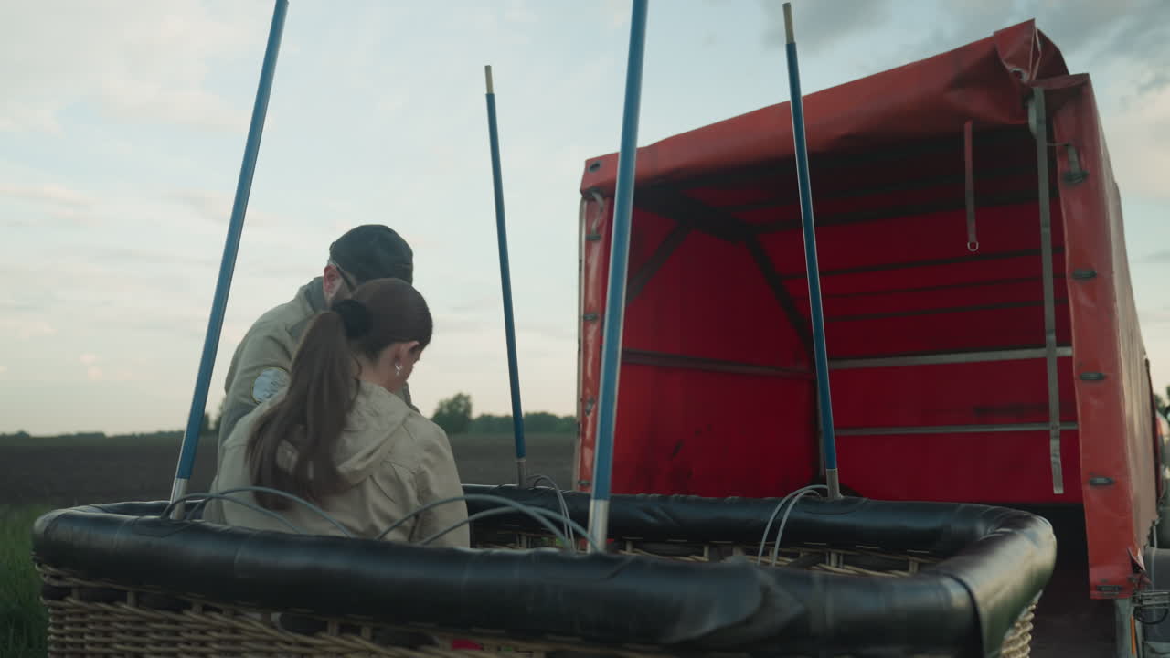 close up of man handing red gas cylinder to woman inside wicker balloon basket on grassy field under cloudy sky, focus on careful transfer motion and safety preparation ritual before flight