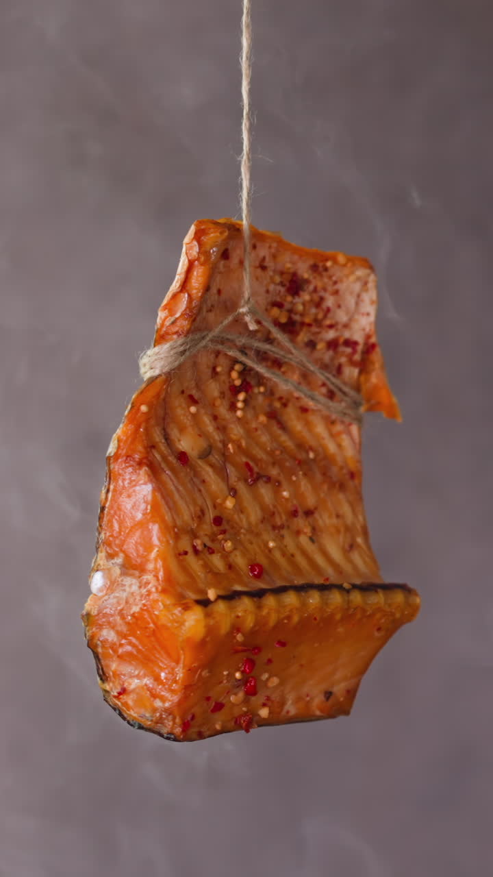 Piece of delicatessen smoked trout fish red meat with spices hangs on rope at rising up white steam on grey background closeup slow motion