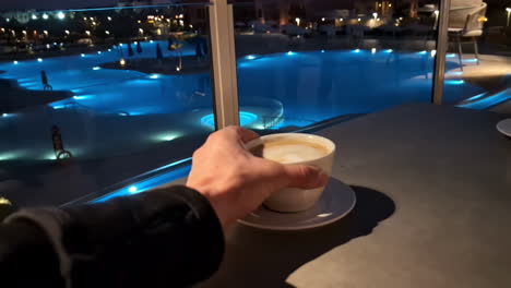 pov of hand picking up coffee from table with luxurious swimming pool backdrop