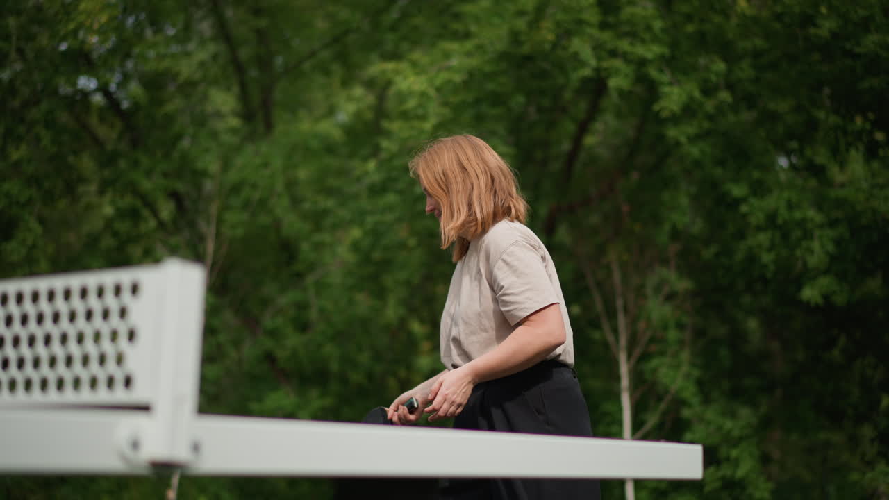 Outdoor Table Tennis Practice With Caucasian Woman Preparing Serve At Park Table, Paddle In Hand, Focused Smile, Casual Clothing, Green Trees Background, Warm Daylight, Candid Motion And Beginner