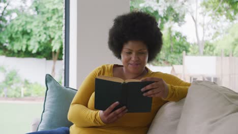 video de una feliz mujer afroamericana de tamaño grande acostada en el sofá y leyendo un libro