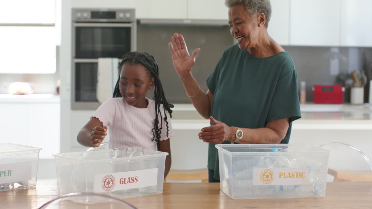 African senior woman guiding youth girl picking, placing jar into GLASS bin in kitchen, high-fiving