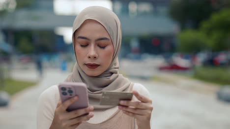 woman in hijab using mobile phone and credit card outdoors