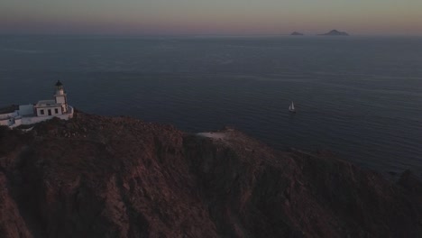 View-of-a-lighthouse,-on-top-of-the-rocky-cliffs-of-Santorini-island,-at-sunset