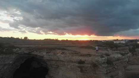 Man-standing-on-rocky-cliff-near-Punta-della-mola-at-sunset,-aerial