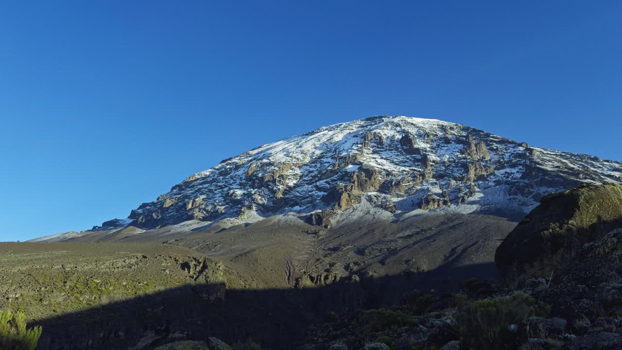 Wide shot shows snow-capped Mount Kilimanjaro rising above a green moorland under a clear blue sky, capturing the mountain’s rugged slopes and sprawling base