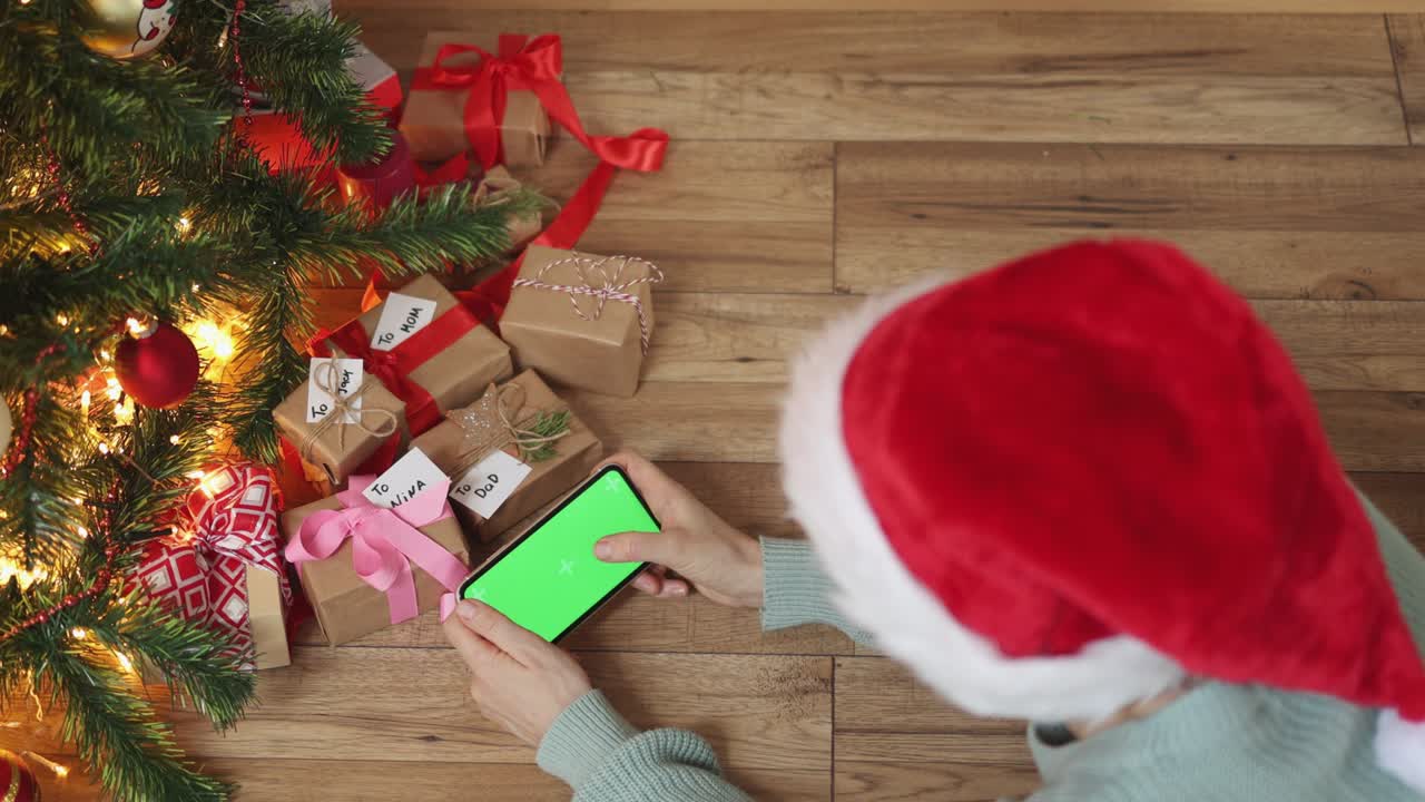 sombrero de navidad rojo femenino manos de teléfono inteligente pantalla verde bajo cajas de regalos de abeto