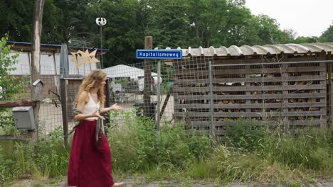mujer caucásica hippie rubia que se detiene para señalar el cartel que dice kapitalisme weg, comunidad anticapitalista, vida sostenible fuera de la red en la aldea ecológica