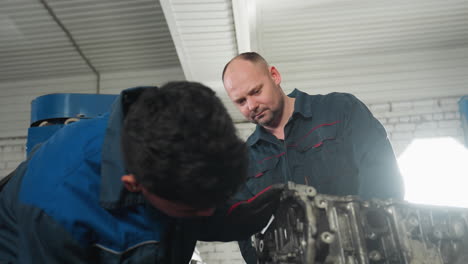 dos ingenieros automotores en un taller, uno intensamente centrado en el ensamblaje de un motor mientras su colega observa atentamente, rodeado de herramientas mecánicas