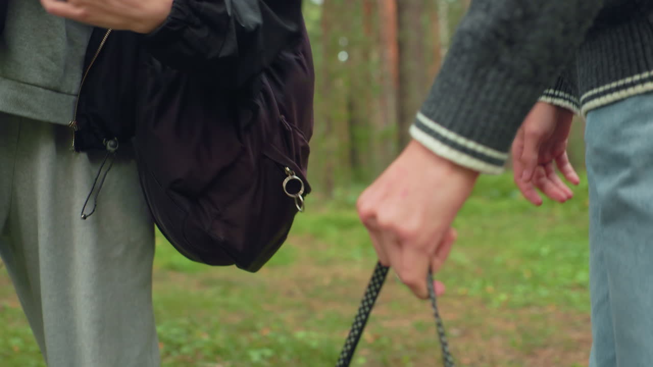 Close up of two companions halting during forest walk, hands relaxed near bags as they prepare to rest or nap, dressed in casual outdoor wear with natural woodland environment in background