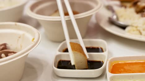chopsticks grasp a tofu slice, dipping it into dark soy sauce beside chili sauce. warm indoor lighting, shallow depth of field, steady close-up shot