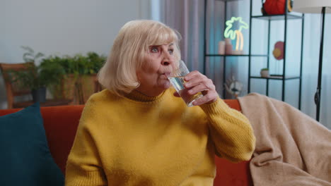 Thirsty-grandmother-woman-sits-at-home-holding-glass-of-natural-aqua-make-sips-drinking-still-water