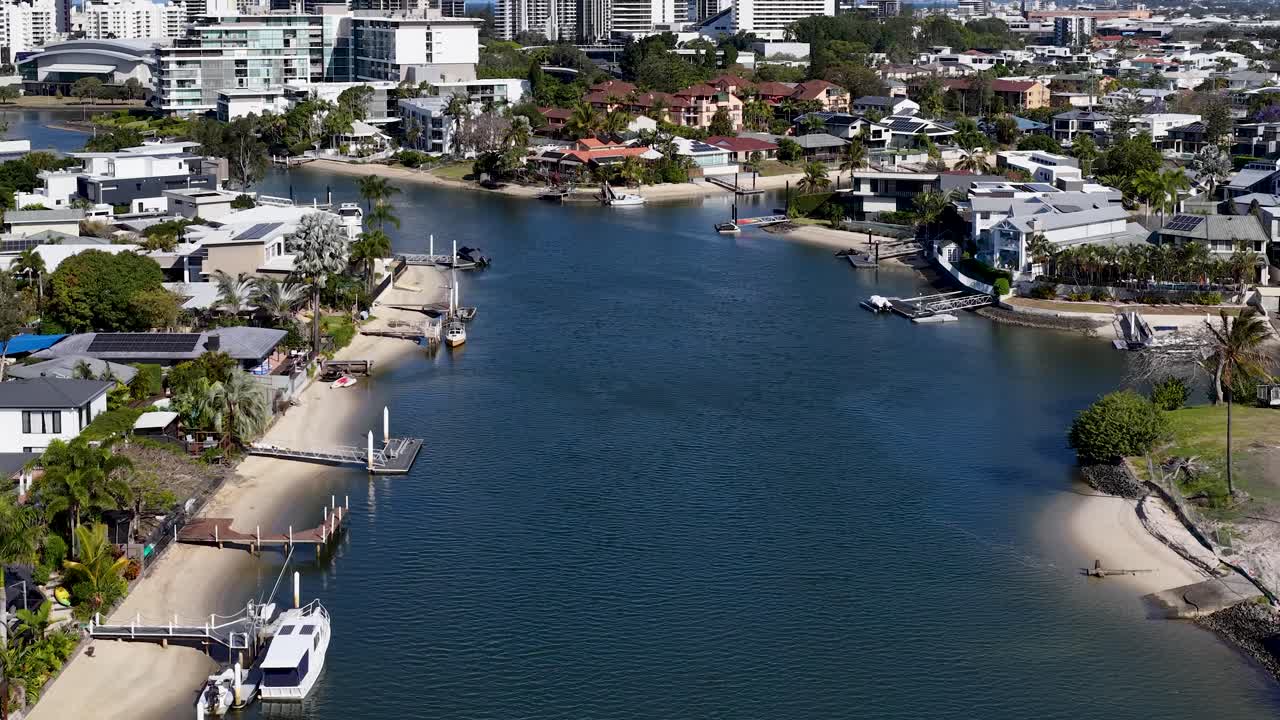 Drone glides above residential canal, modern houses, private docks, and boats in bright daylight