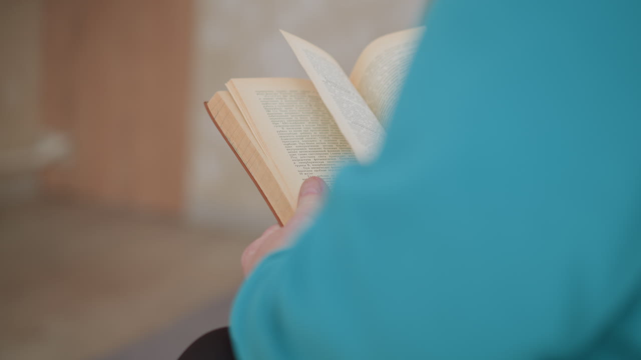 Close-up shot of lonely pensioner holding brown book in hands, reading in cozy home environment. Elderly woman in green teal sweater focuses on book, peaceful indoor scene with soft lighting