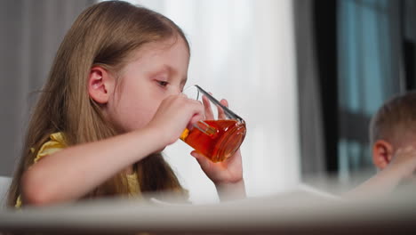 close up of little child seated drinking fruit wine from transparent glass cup with kid brother sitting next to her at dining table in cozy room as soft daylight filters through curtains