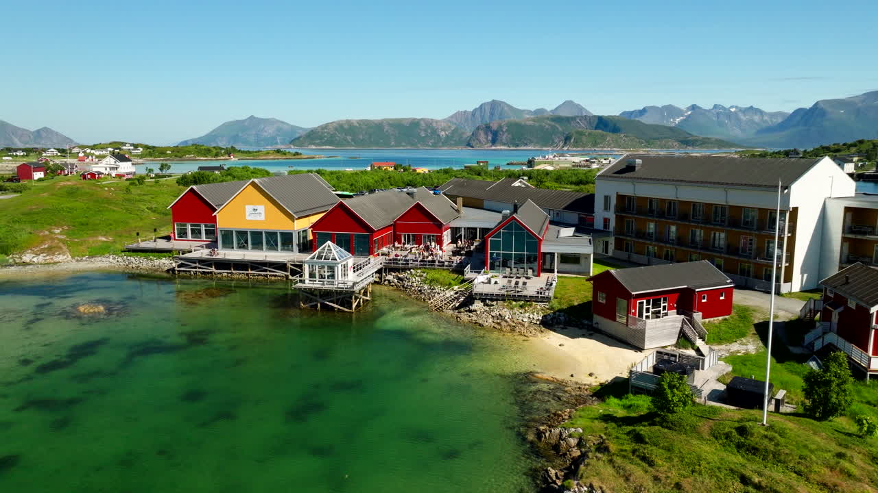 Sommarøy Arctic Hotel, Troms, Norge, fargerike bygninger på stylter over turkis vann, fjell i bakgrunnen under solfylt sommerhimmel, dronefoto i bane