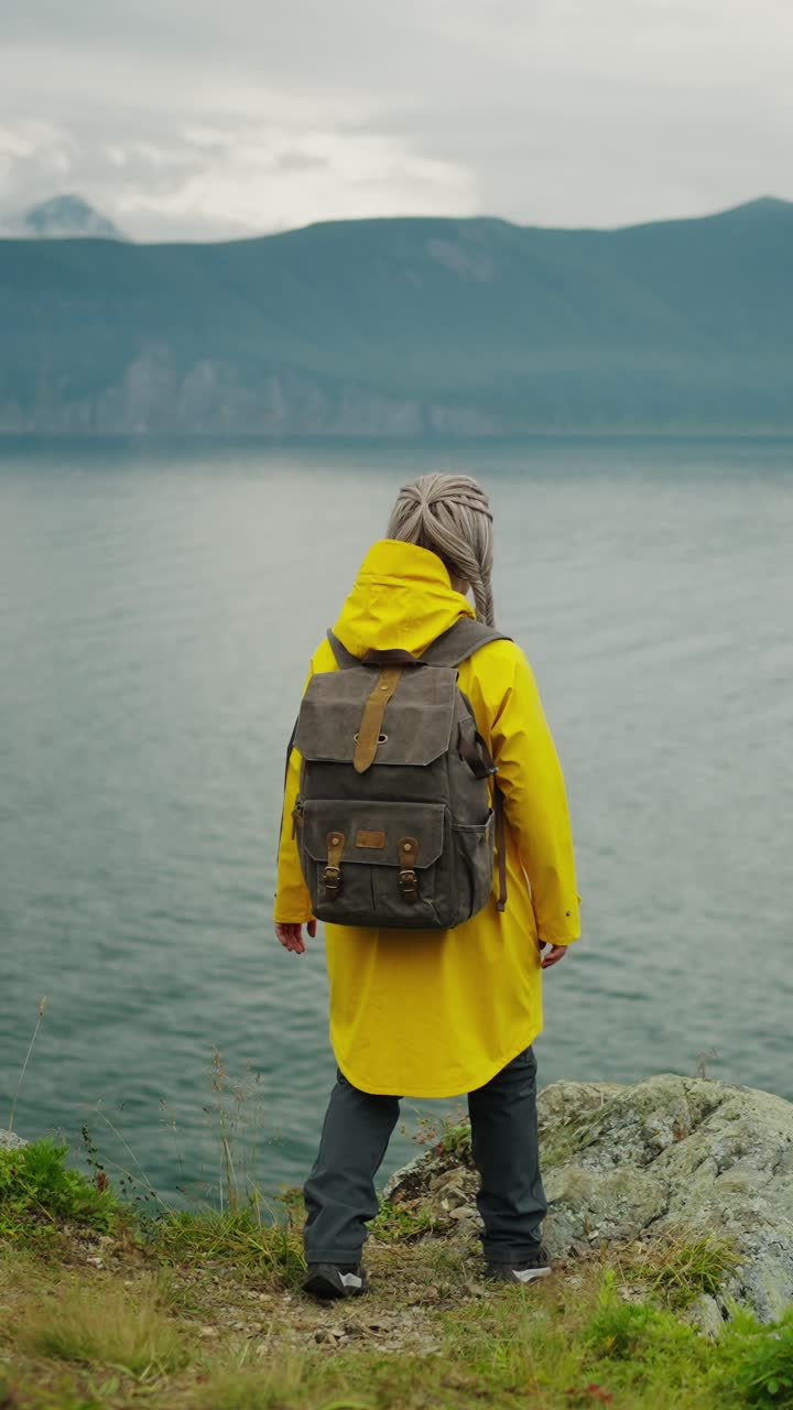 Donna in impermeabile giallo con zaino in piedi accanto a un lago e montagne