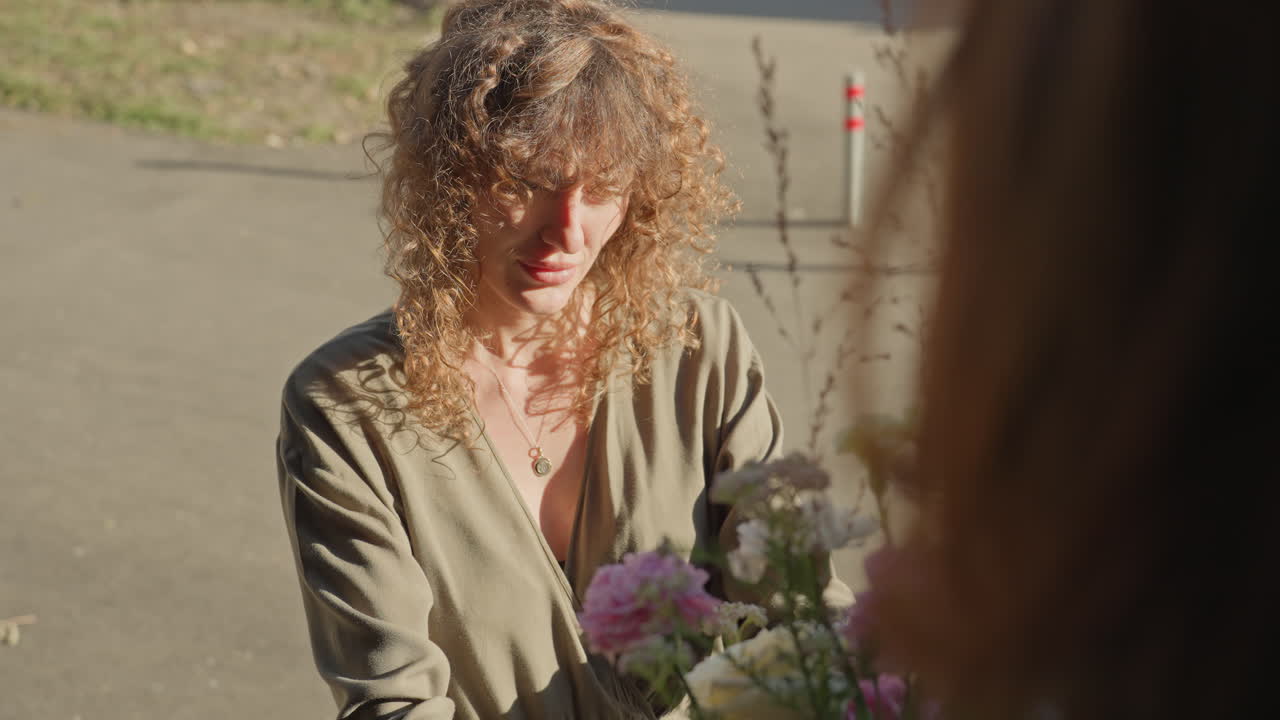 Outdoor florist handing bouquet to customer, white curly woman exchanging sunlit pastel arrangement, hands adjusting roses and peonies, soft morning light, market stall mood, delicate petals