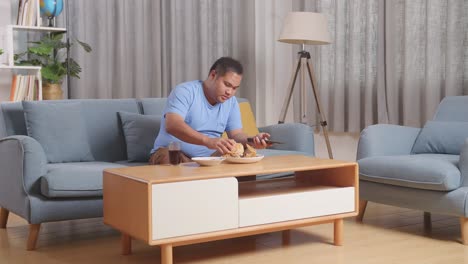 full body of a fat asian man eating fast food having hamburger while watching tv on a sofa in the living room at home