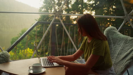 woman writes last lines finishing morning work on laptop in glamping house. busy woman alone sits in armchair with cup of coffee on low table