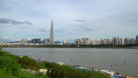 Gente-Disfrutando-Del-Río-Hangang-Recreativo---Windsurf,-Paddle-Surf-Y-Kayak-Con-El-Horizonte-De-La-Ciudad-De-Seúl-En-El-Fondo,-Lotter-World-Tower