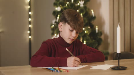 Boy-In-Red-Turtleneck-Writing-A-Letter-Of-Wishes-Sitting-At-A-Table-In-A-Room-Decorated-With-A-Christmas-Tree