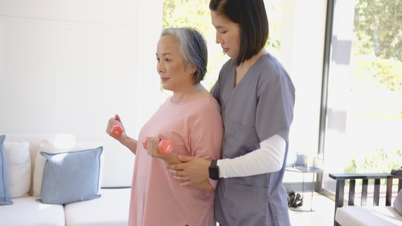 Assisting senior asian woman with dumbbells, physical therapist guiding exercise at home