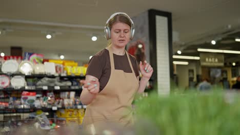 Confident-girl-supermarket-worker-in-a-brown-T-shirt-and-apron-in-wireless-headphones-listening-to-music-and-arranging-goods-on-the-counter-in-a-supermarket