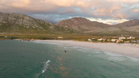 Single-kitesurfer-riding-towards-the-beach-in-Pringles-Bay,-South-Africa-during-sunset,-drone-shot