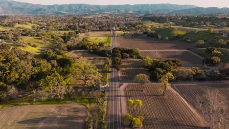 Drone-Over-Farm-Fields-in-Santa-Ynez-California,-Cars-Driving-Down-Wine-Country-Road-at-Golden-Hour-with-Mountains-on-Horizon