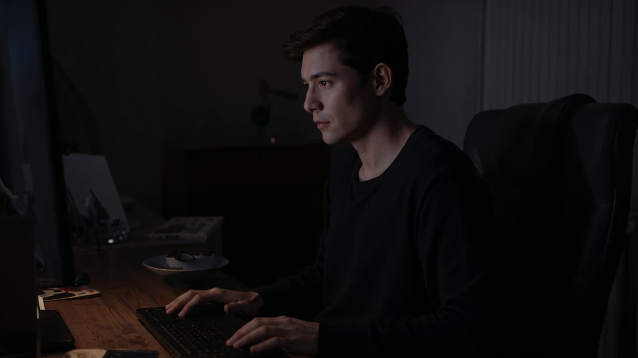 Typing man in dark shirt at dim home workspace for work, reaching monitor and adjusting keyboard