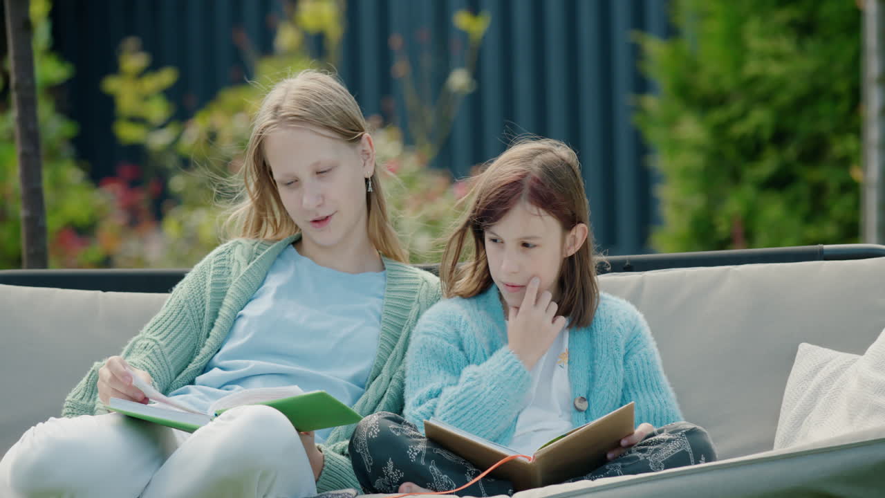 Due ragazze adolescenti leggono libri, sedute su un'altalena da giardino nel cortile sul retro della casa.