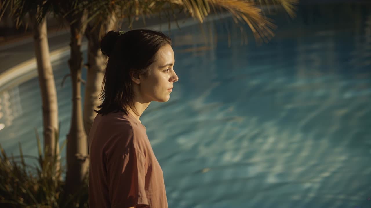 Standing woman in mauve tee lifting chin poolside, sunlight warming face, watching rippling water