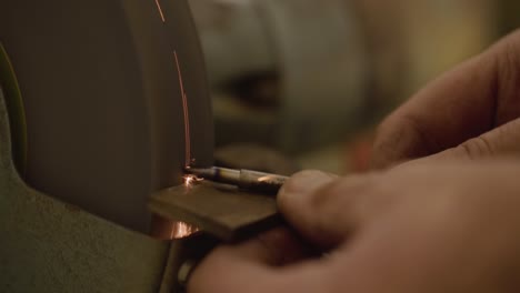 Blacksmith-using-grinding-machine-with-whetstone-to-form-a-metal-tool,-sparks-flying-towards-the-camera-in-slow-motion-close-up-macro