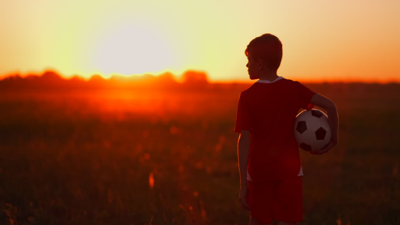 el niño está en el campo al atardecer en la hierba con una pelota de fútbol. soñando con una carrera futbolística. el concepto de éxito en el deporte.