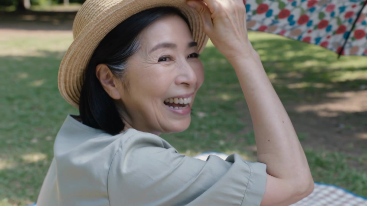 カメラが引いていくと、薄手のシャツを着た年配の女性が笑いながら麦わら帽子を抑えている。公園でのピクニックの光景。