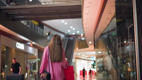 vista trasera de una mujer con un vestido rosa sosteniendo una taza de café y caminando a través de un centro comercial brillantemente iluminado, el fondo borroso presenta tiendas, luces y efecto bokeh, mientras la gente camina en la distancia