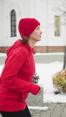 side view of woman jogging along snowy park pathway in red hoodie and black leggings by benches and white building surrounded by serene frosty trees bushes and cold atmosphere conveying cold fitness