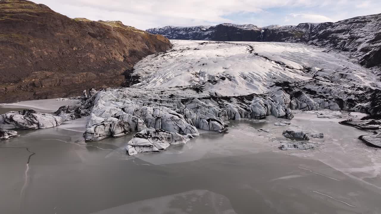 Aerial view of the glacial tongue of Sólheimajökull, Iceland, meeting a milky glacial lagoon. Rugged landscape showing ice formations and meltwater.