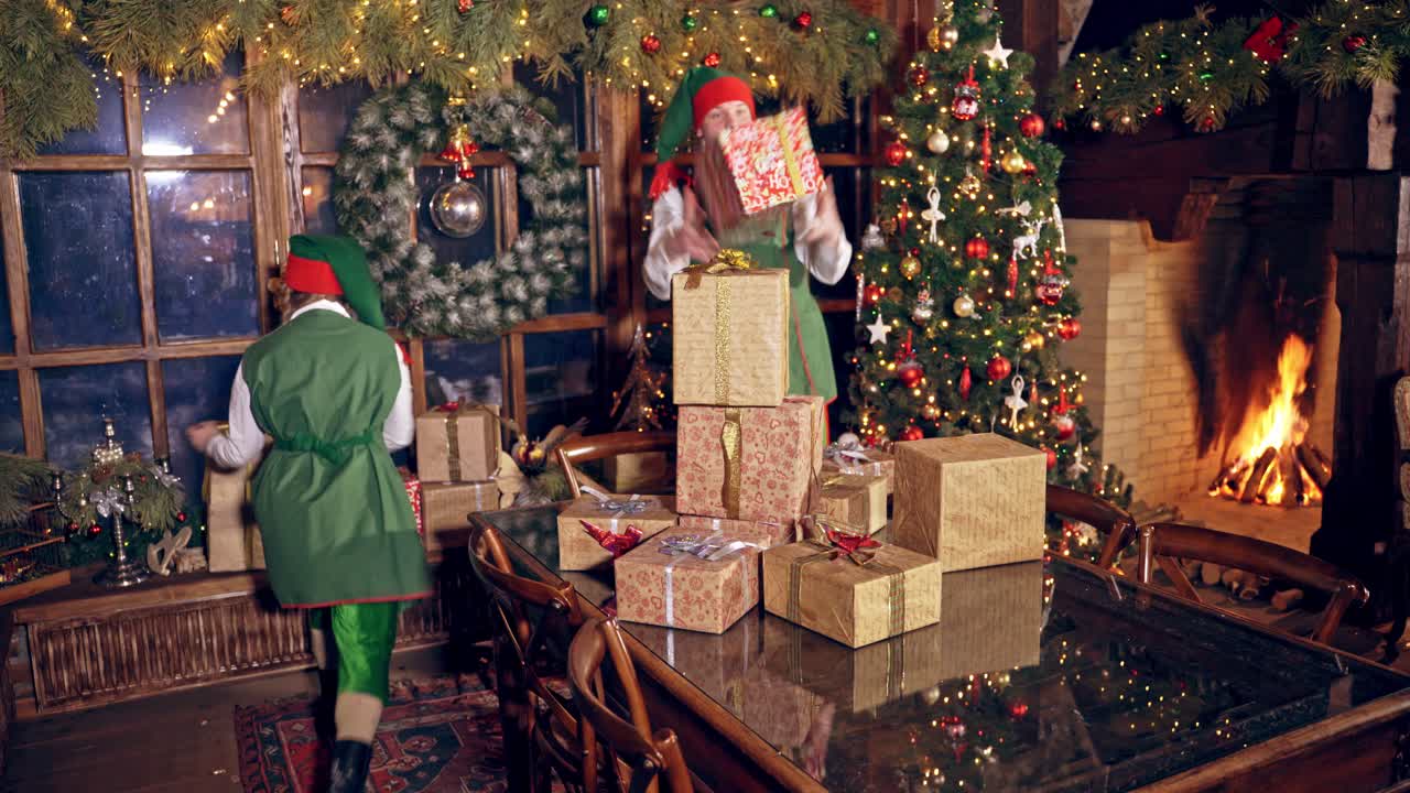 Happy elves with many presents indoors. Christmas decorated room and elves in green costumes putting packed gifts on the table. Christmas time.