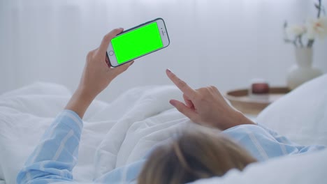침대 위에 휴대 전화를 가진 젊은 여성, 어깨 뷰. 기술 중독. 녹색 화면 전화. 침실에서 배를 만지고 스마트폰을 사용하는 젊은 여성