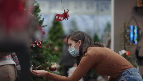 a woman in a protective mask chooses christmas decorations. preparing for christmas in the coronavirus pandemic. christmas tree toys and garlands