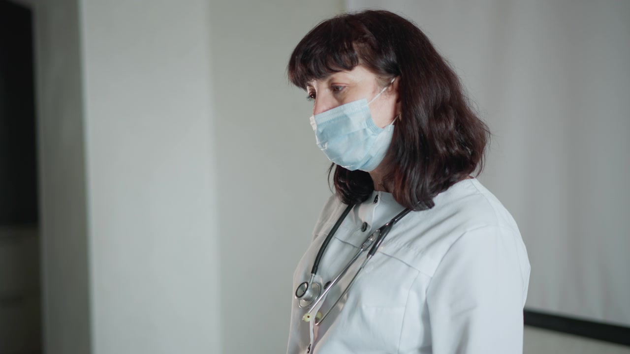 Inquadratura ampia di un'infermiera che tiene una valigetta medica, operatrice sanitaria bianca con mascherina e stetoscopio, che entra nel soggiorno, con una chiara cassetta di pronto soccorso in mano, pronta per la visita al paziente, precauzioni covidsafe