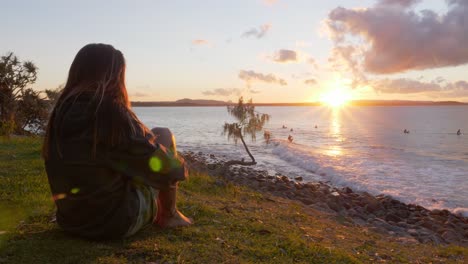 Mujer-Sentada-En-El-Césped-Observa-A-Los-Surfistas-Durante-La-Puesta-De-Sol-En-El-Parque-Nacional-De-Noosa-En-Australia