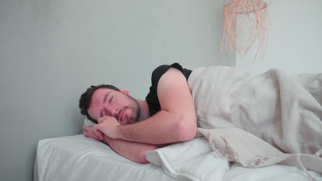 hombre dormido, blanco caucásico con barba, se despierta por la mañana, se estira cansado, abre los ojos, se levanta relajado en el interior del dormitorio