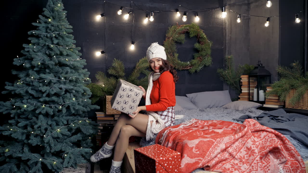 Positive young woman with gift box. Attractive girl in knitted cap and scarf sitting on bed near the Christmas tree with a big present and throwing it happily. Happy New Year.