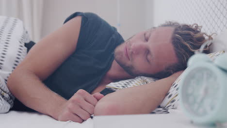 man asleep in bed reaches out to turn off alarm clock on bedside table