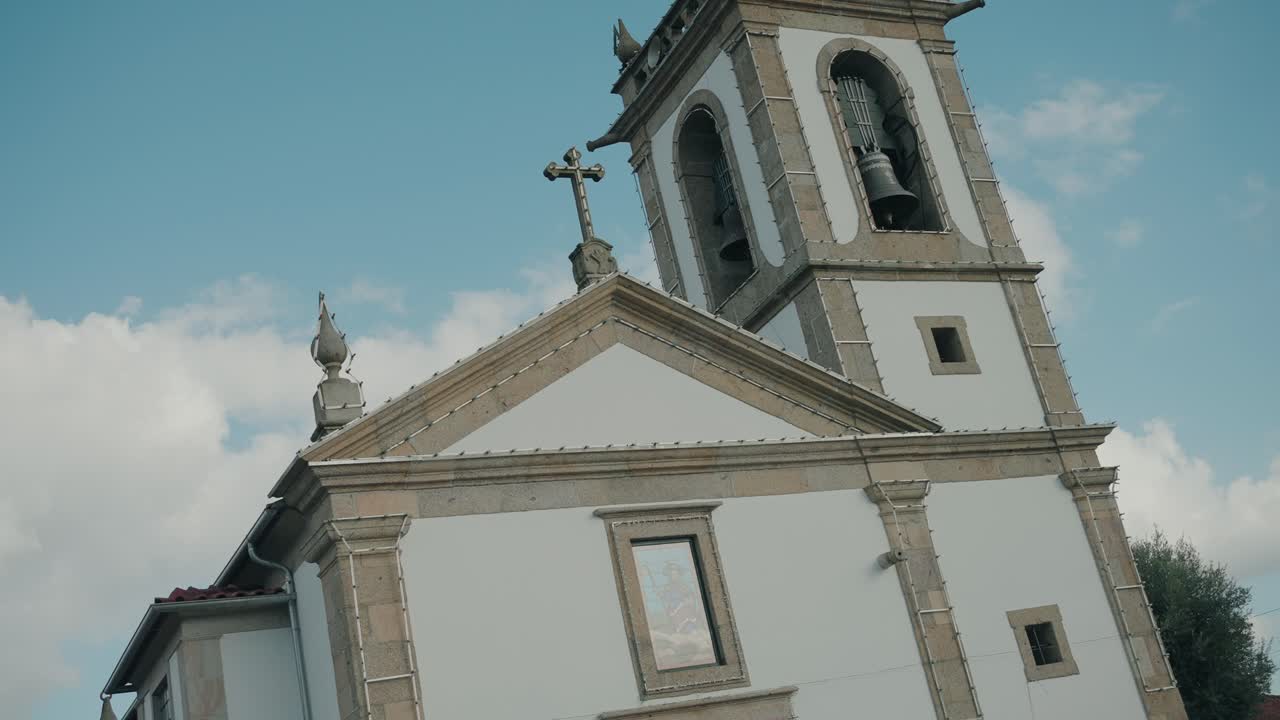 Vista dal basso del campanile bianco di una chiesa barocca con orologio, campane e croce contro il cielo blu