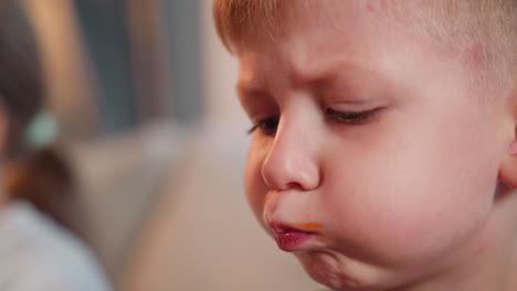 close up of young boy with cheeks puffed out savoring toasted bread with sauce stain on lips as he licks mouth beside blurred figure of girl with ponytail seated near him