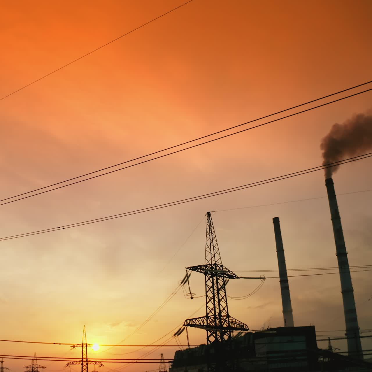 Pipe with dark smoke near the transmission lines. High-voltage power towers next to the harmful manufacture against the epic sky of setting sun. Motion top down